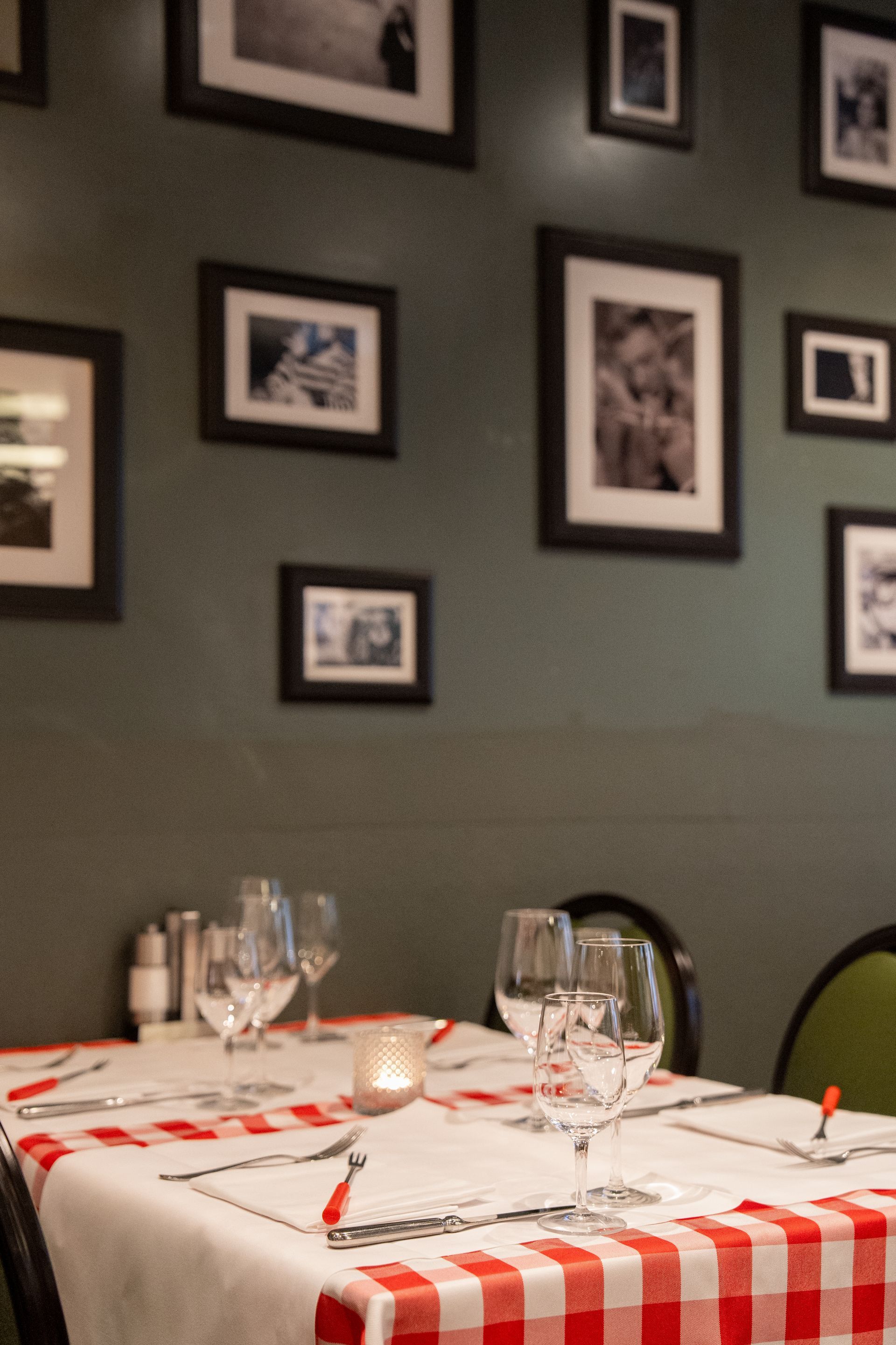Restauranttisch mit weißer Tischdecke, Weingläsern und gerahmten Fotos an einer dunkelgrünen Wand