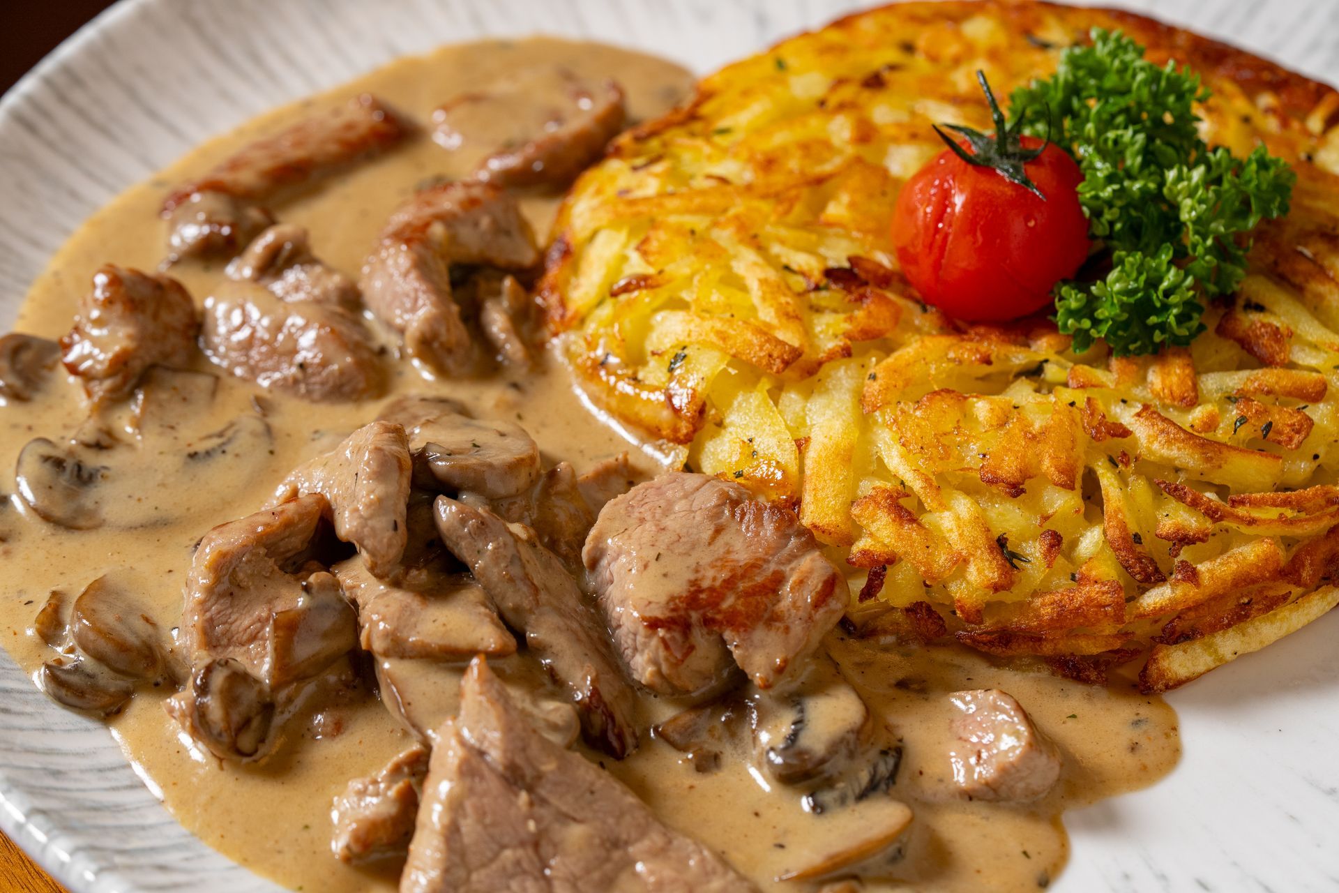 Rindfleisch Stroganoff mit cremiger Pilzsoße und goldbraunen Rösti, garniert mit Petersilie und einer Kirschtomate.