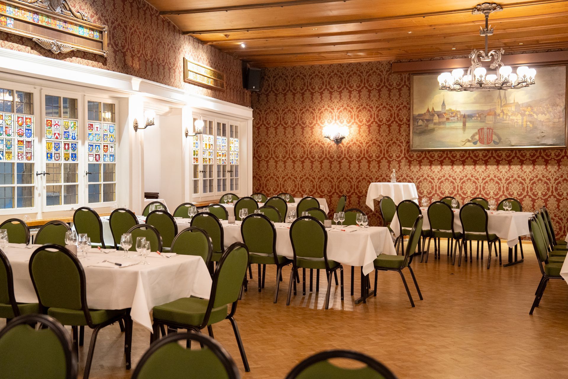 Banquet hall set with long white-draped tables, folded napkins, and chandeliers under a wood-paneled ceiling