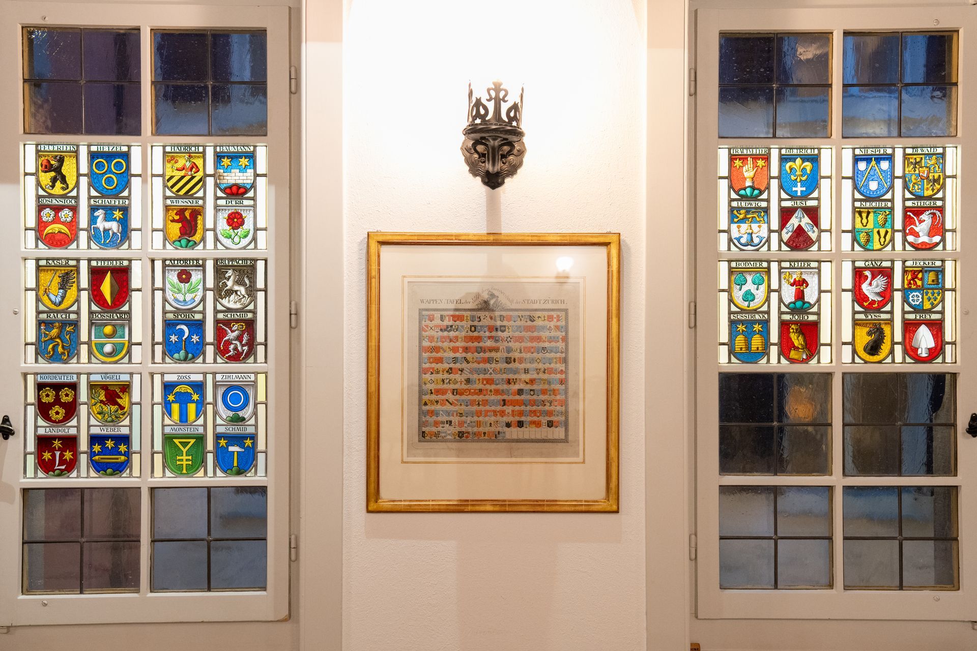 Buntglasfenster flankieren eine gerahmte Gedenktafel an einer weißen Wand unter einer dekorativen Wandleuchte.