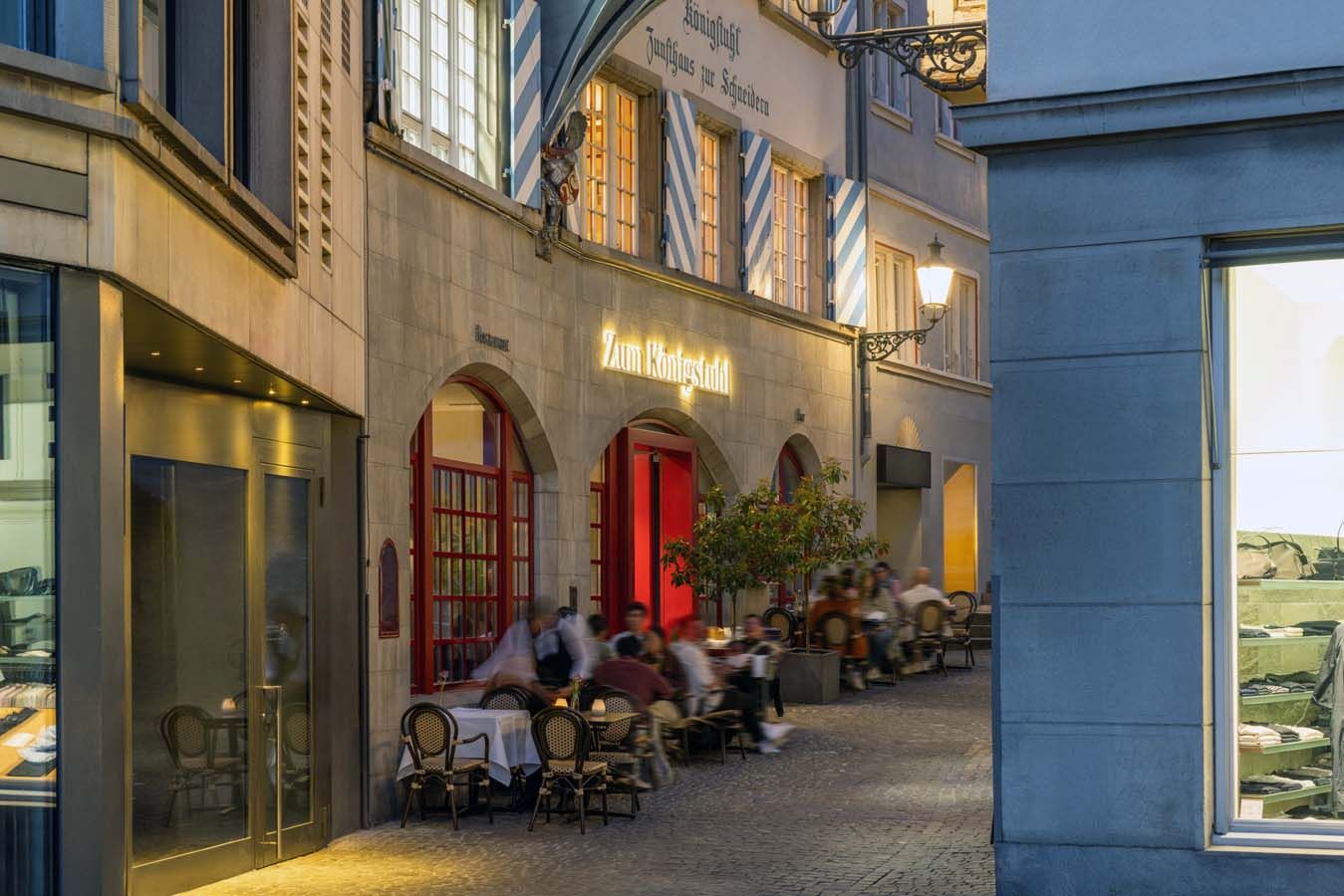 Menschen speisen abends an Tischen in Straßencafés entlang einer warm beleuchteten Straße.