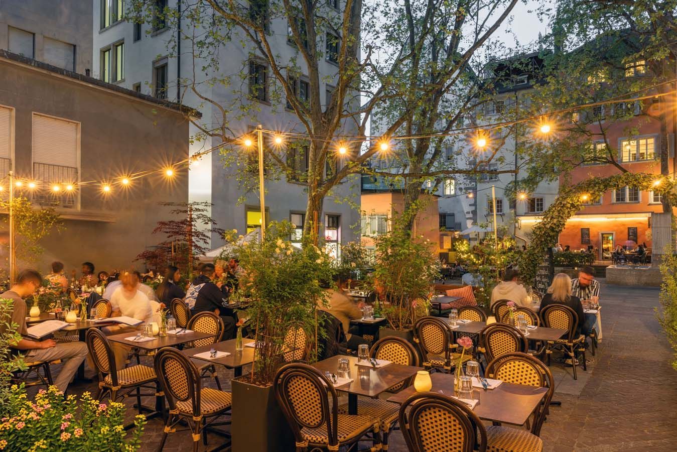 Terrasse eines Stadtcafés in der Abenddämmerung mit Lichterketten, vielen Gästen und Topfpflanzen.