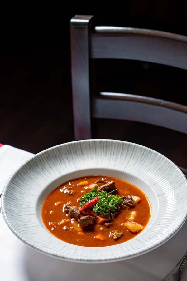 Eine weiße Schüssel mit roter Suppe und Garnitur steht auf einem Tisch neben einem Holzstuhl.