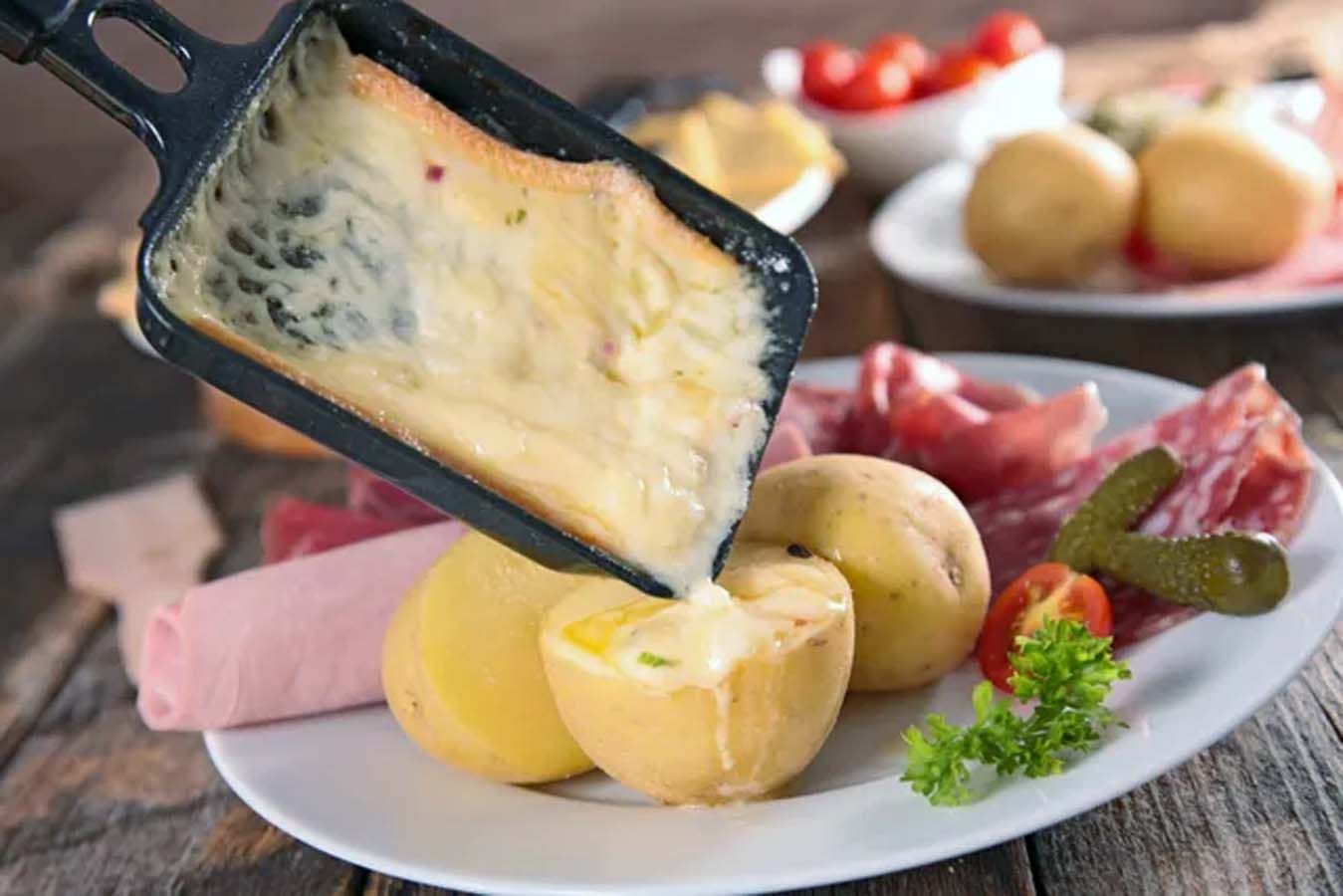 Geschmolzener Raclettekäse wird über Kartoffeln gestrichen, dazu Gurken und Schinken auf einem Teller.