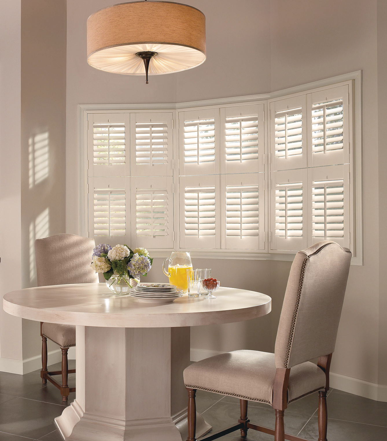 Bright dining nook with round table, beige chairs, white shutters, and breakfast setting by the window