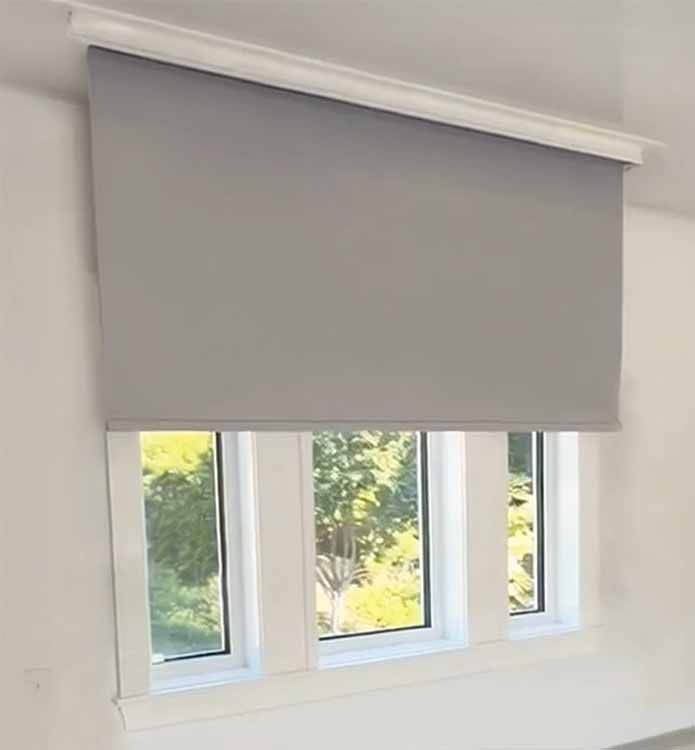 Gray roller blind lowered over a window, with three panes and trees visible outside.