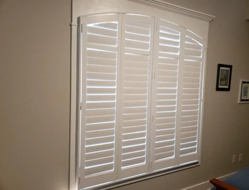 White plantation shutters on a beige wall beside framed art in a room.