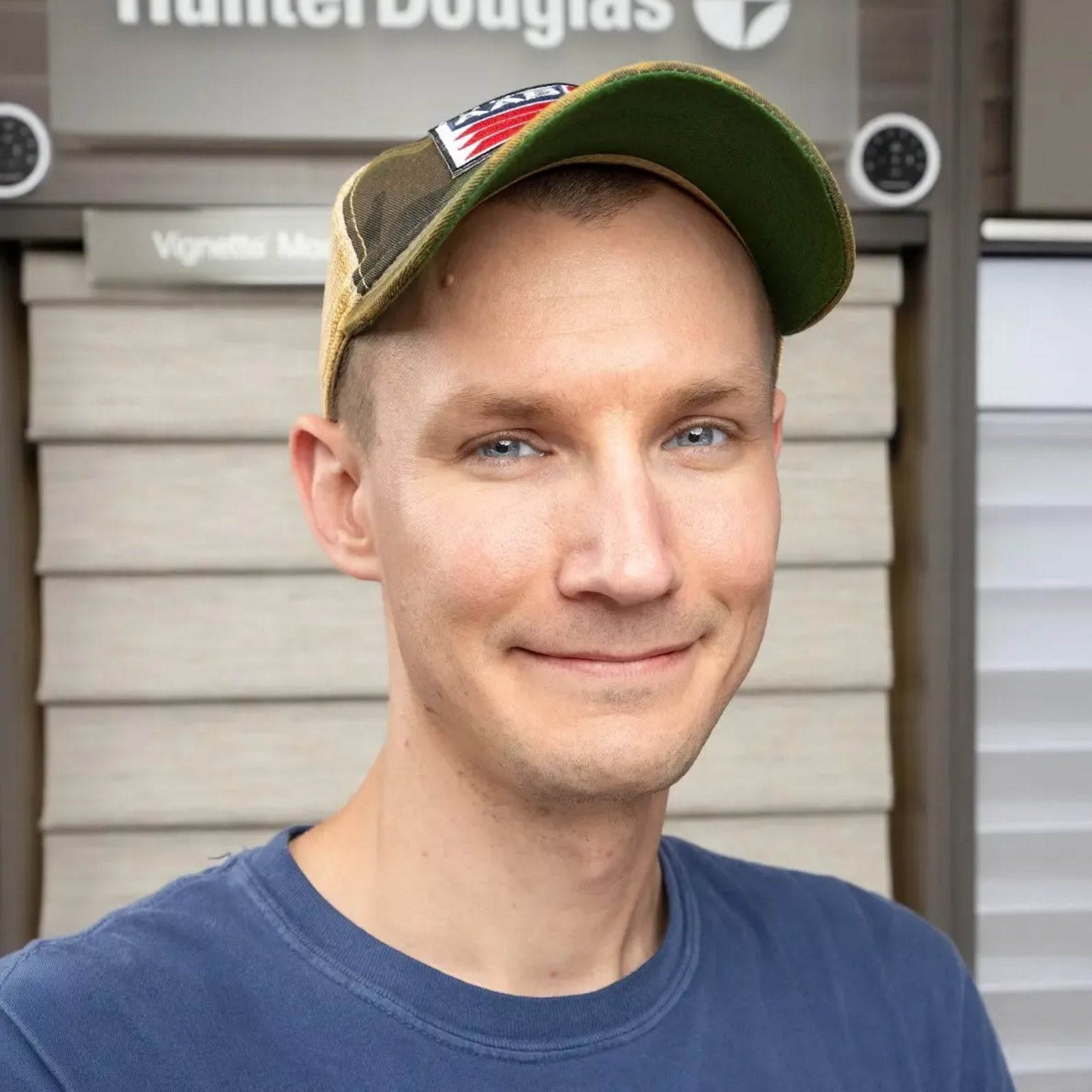 Man in a green cap and blue shirt smiling in front of a Hunter Douglas storefront