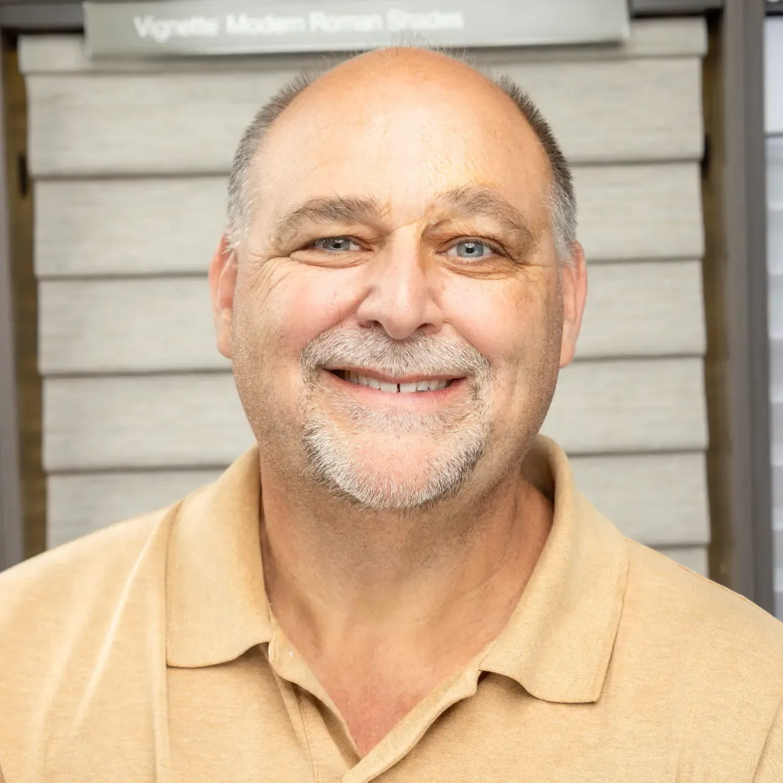 Smiling man in a tan polo shirt standing in front of a gray wall.