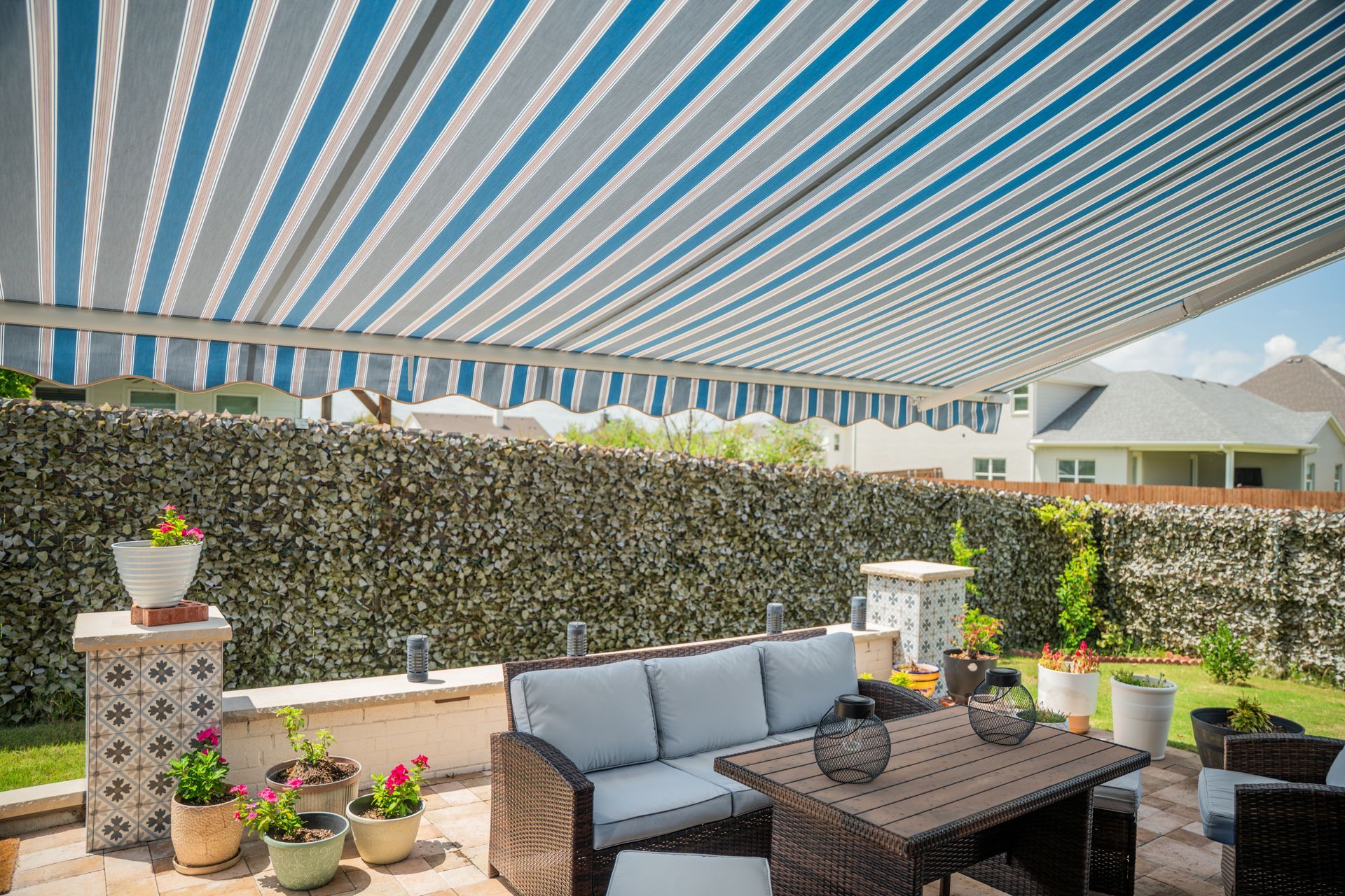 Backyard patio with striped awning, wicker sofa, table, potted plants, and a hedge fence