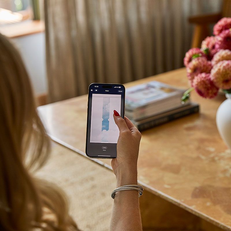 Person holding a smartphone over a table, viewing a photo app beside flowers and books.