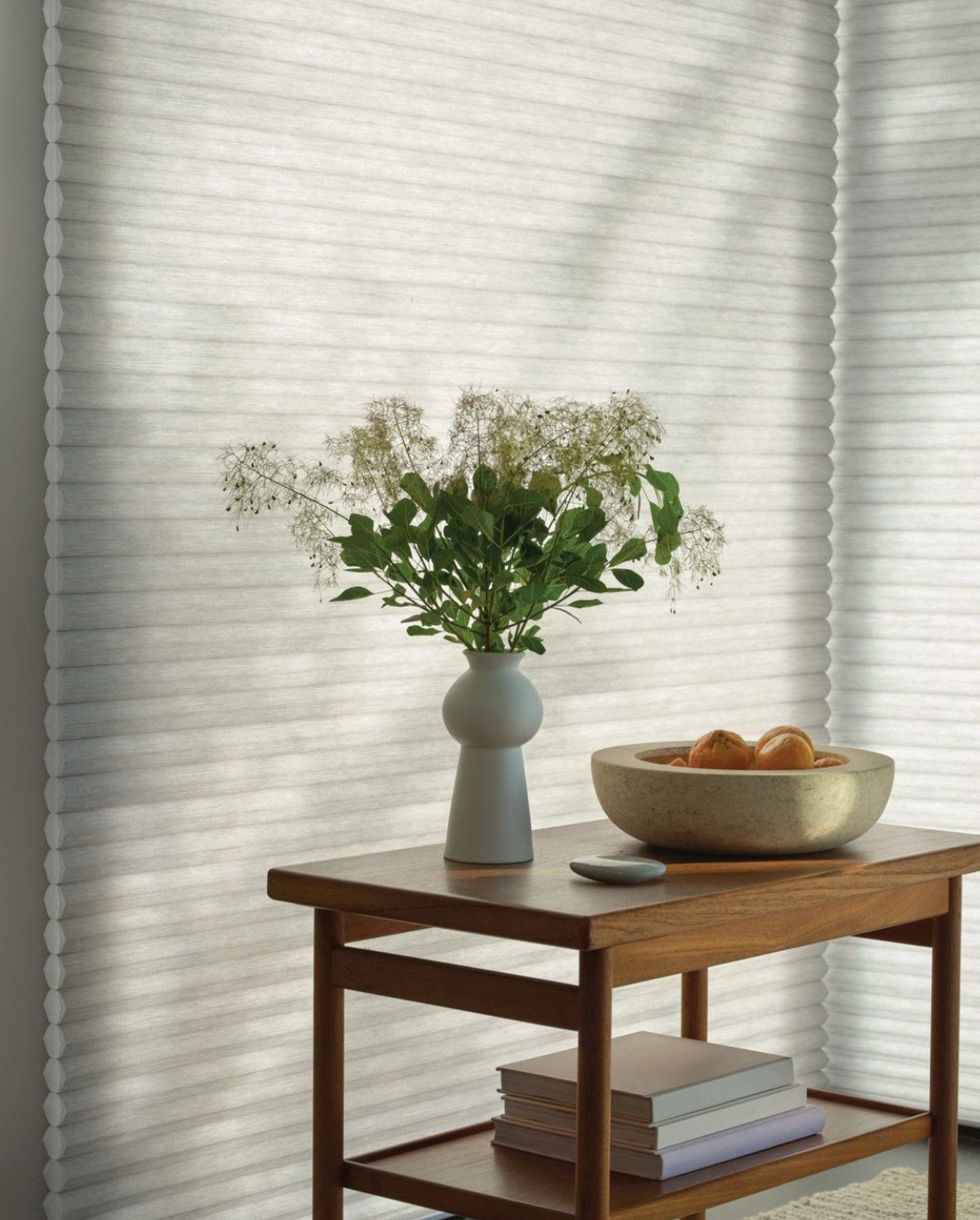 Vase of white flowers on a wooden table beside a bowl of fruit in a sunlit room