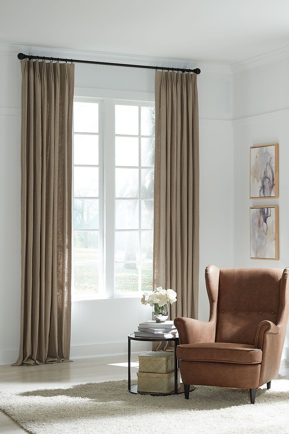 Bright living room with tall windows, beige curtains, and a brown armchair beside a small table and flowers.