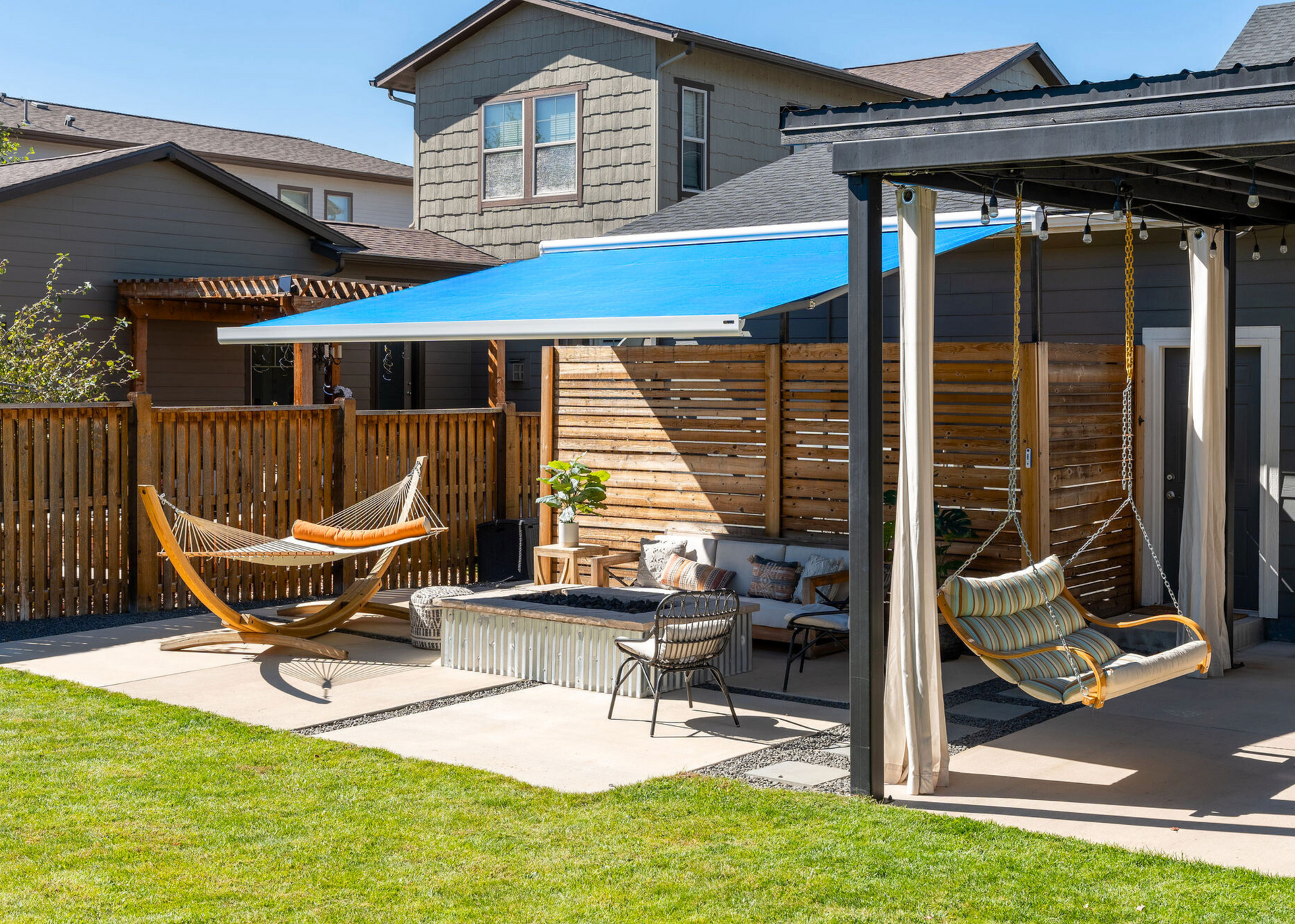 Backyard patio with blue canopy, hammock chairs, fire pit, and wooden fence behind a lawn