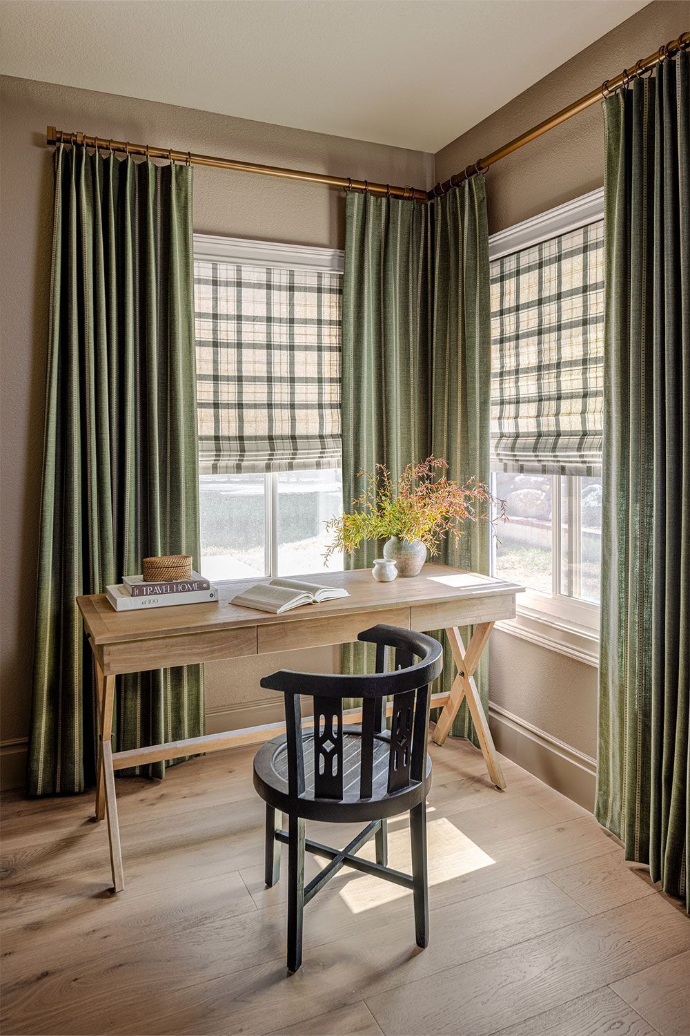 A cozy corner desk by tall windows with green curtains, a black chair, and flowers on the table.