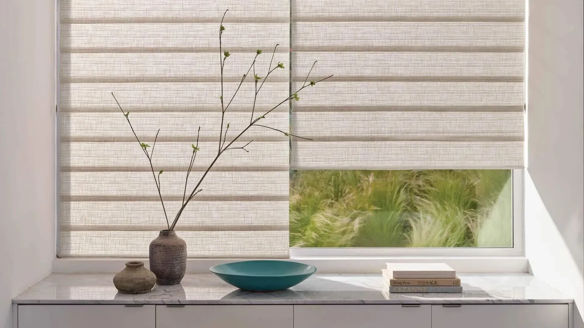 Minimalist window ledge with beige blinds, a vase of branches, bowl, and stacked books.