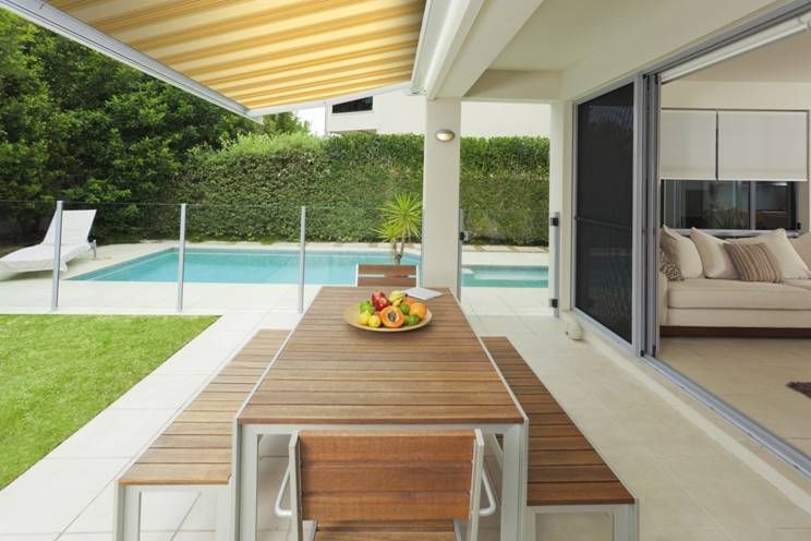 Covered patio with wooden dining table, pool, and garden view beside an open sliding door.
