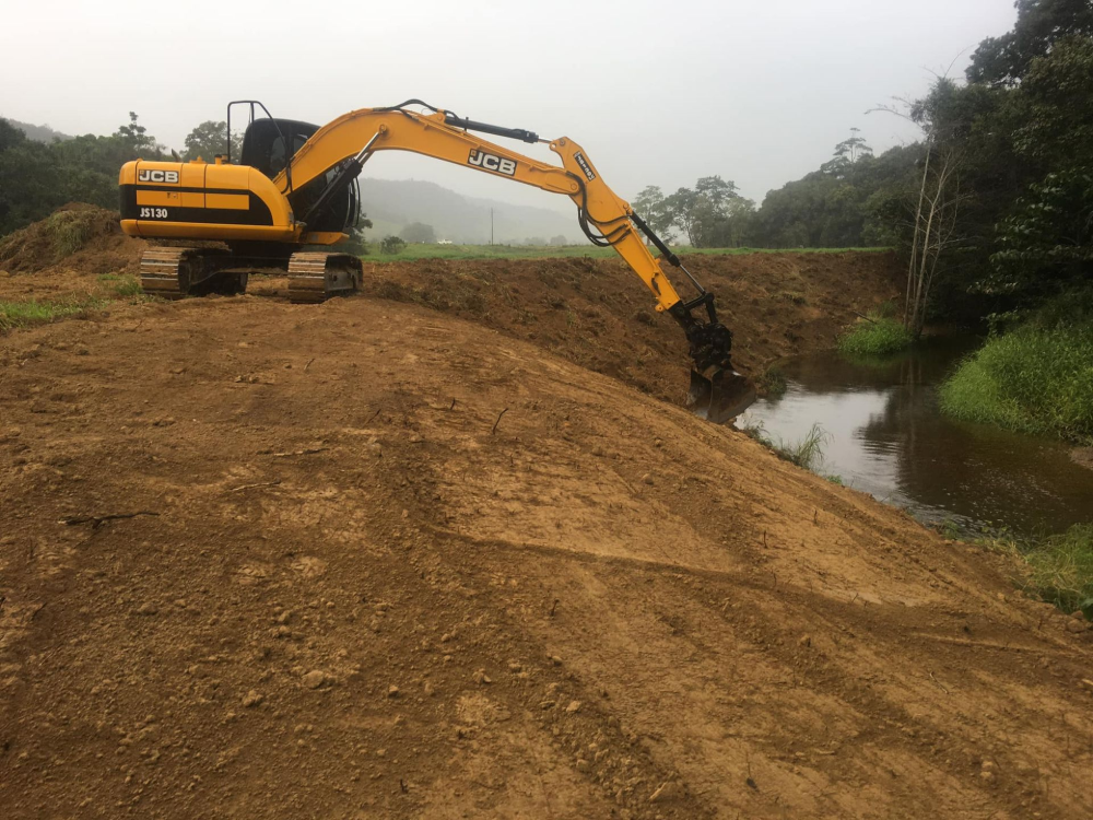 Yellow excavator digging at a riverbank, moving dirt. Green trees and overcast sky in background.— Macca's Dynamic Earthworks in Tolga, QLD