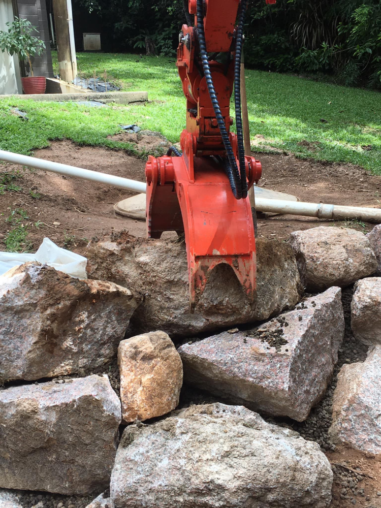 An orange excavator claw gripping a rock in front of a stone retaining wall.— Macca's Dynamic Earthworks in Tolga, QLD