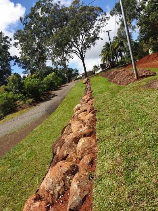 A Rock Wall Lines a Grassy Hillside Alongside a Road with Trees and Blue Sky — Macca's Dynamic Earthworks in Tolga, QLD