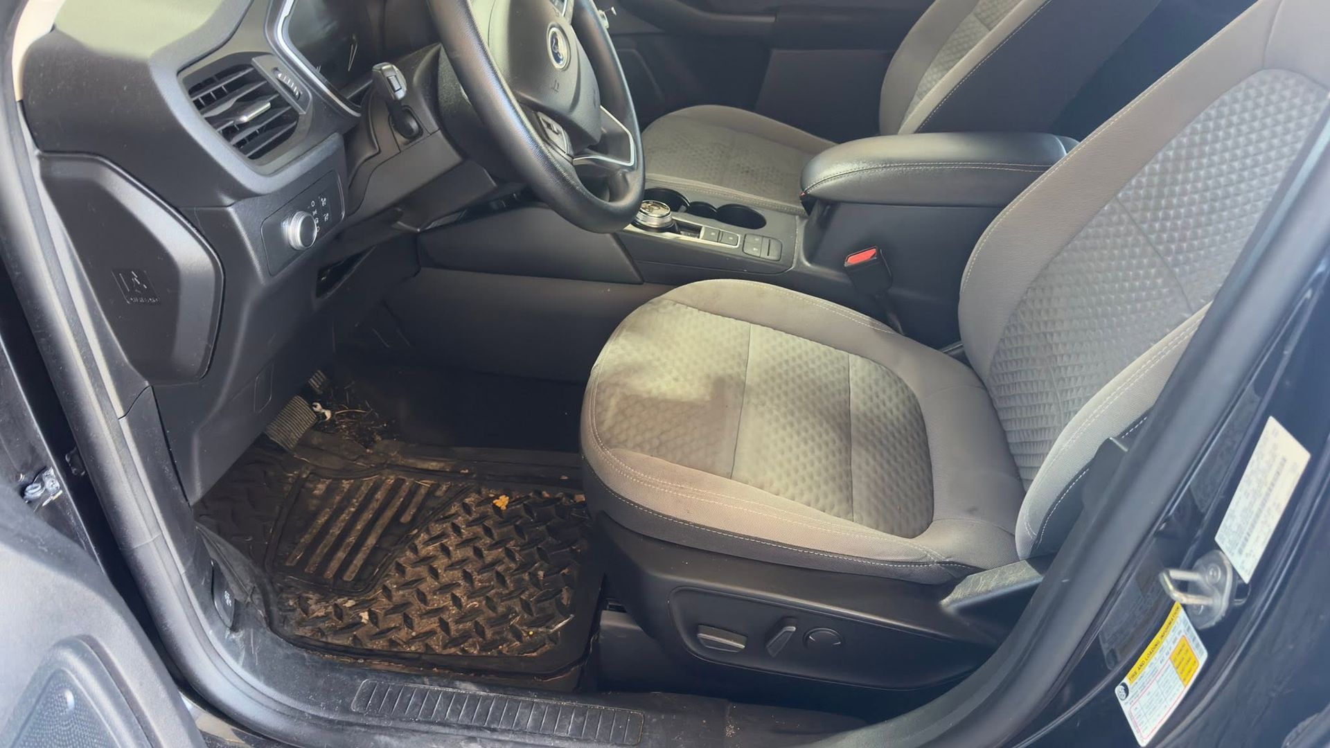 Car interior with front seats, steering wheel, and rubber floor mat visible from the open driver-side door