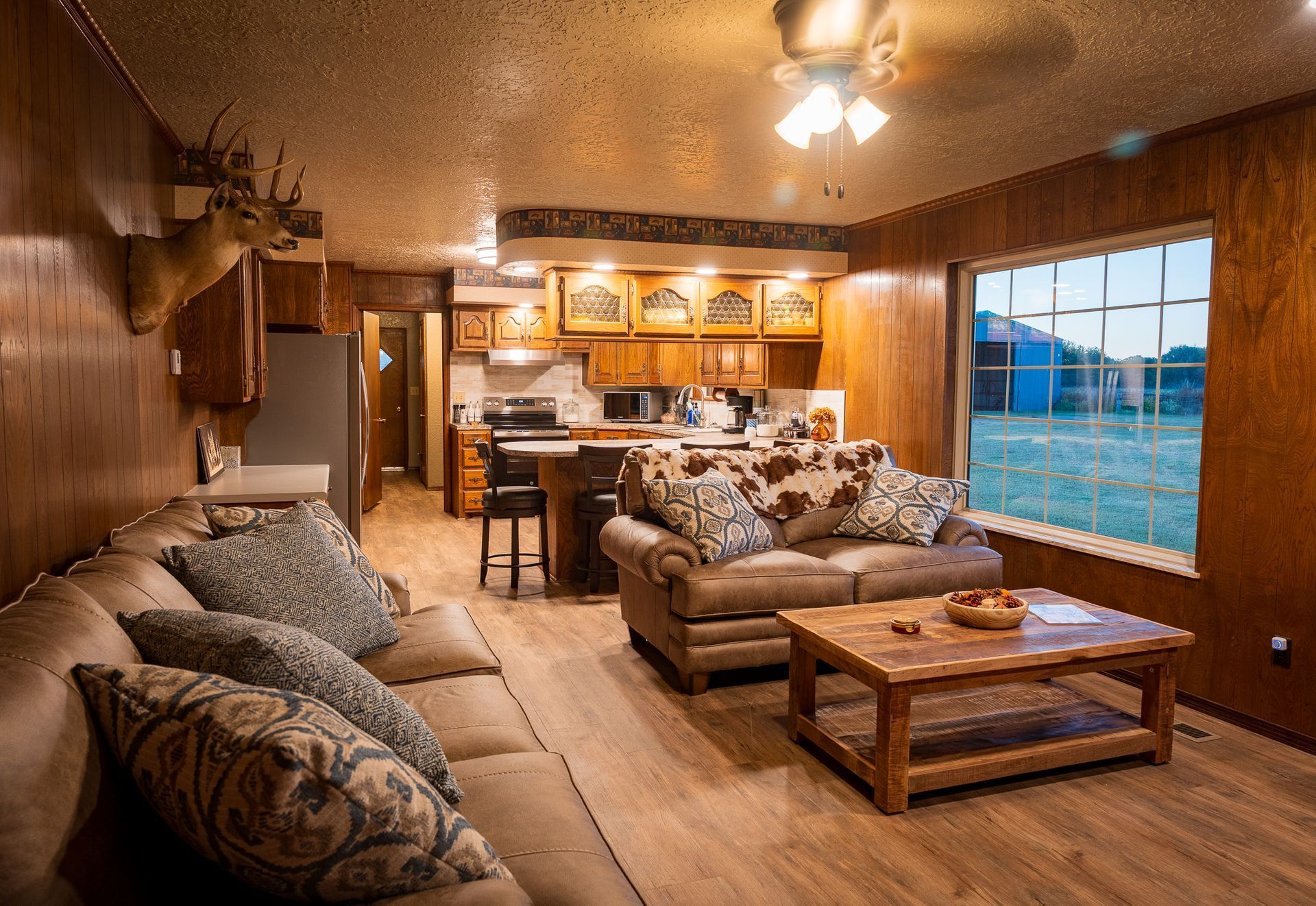 Wood-paneled living room with brown sofas, coffee table, and kitchen visible by large window at sunset