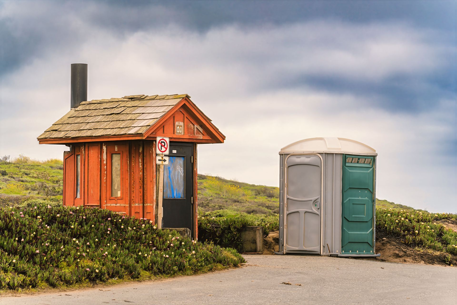Small orange hut and green portable toilet beside a road on a grassy coastal hillside.