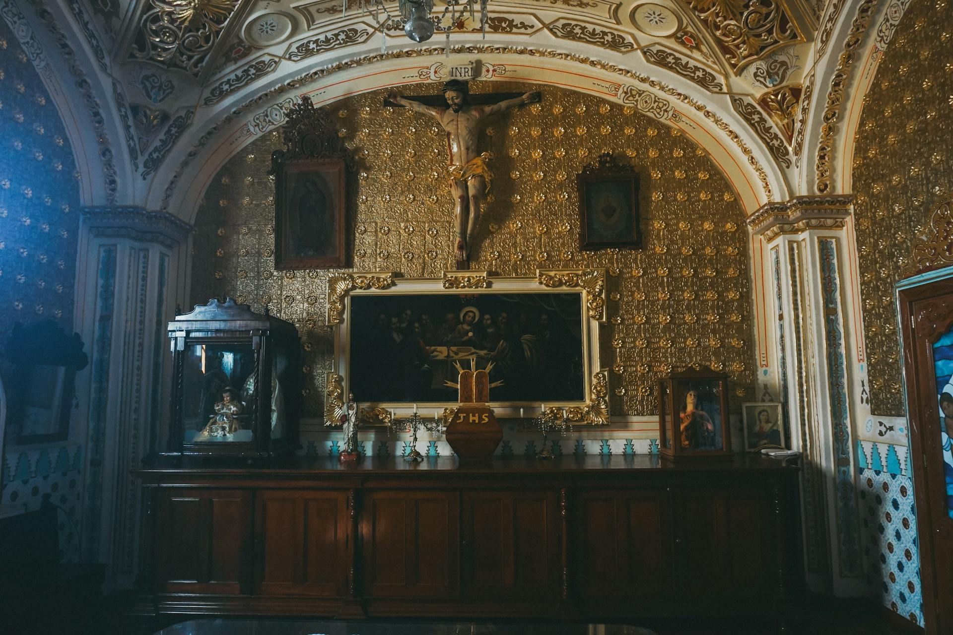 An ornate chapel with gold-patterned walls, a wooden altar, a crucifix, and framed artwork under a vaulted ceiling.