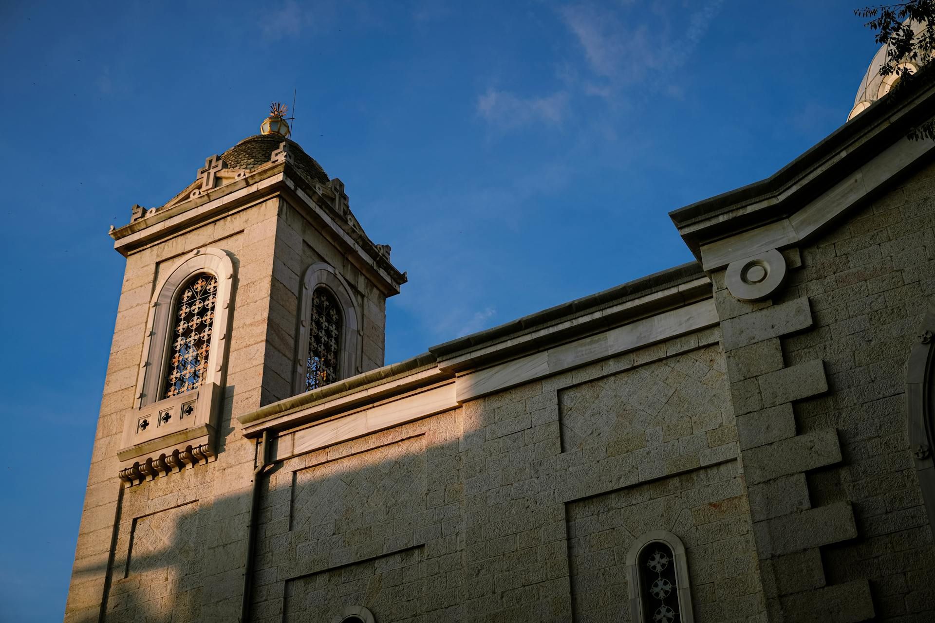 Low-angle view of a tan stone church tower and exterior wall against a clear blue sky during sunset.