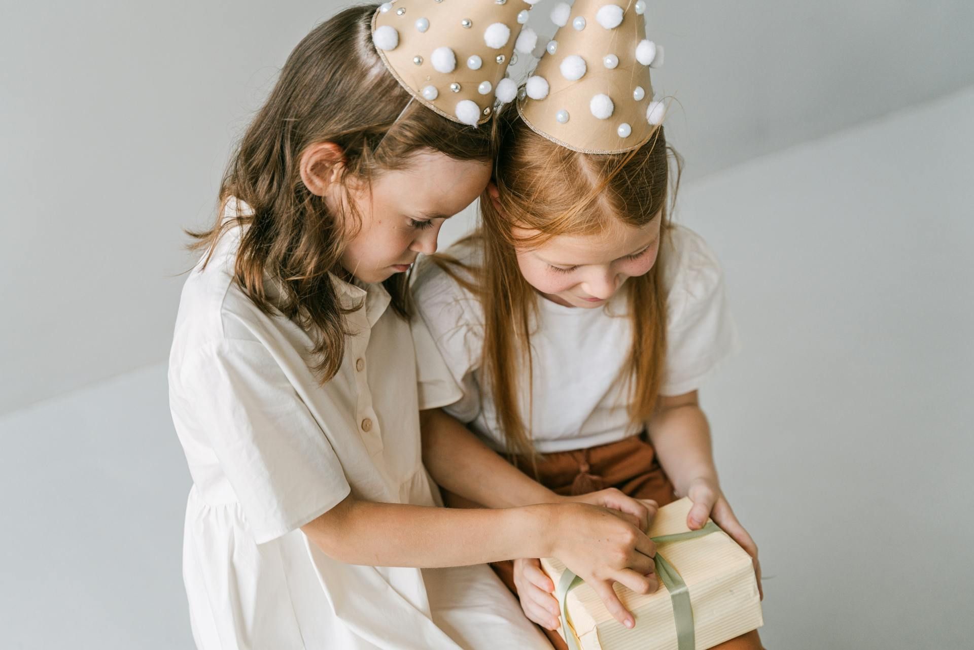 Two children in party hats sit together and focus on unwrapping a small gift wrapped in tan paper and green ribbon.