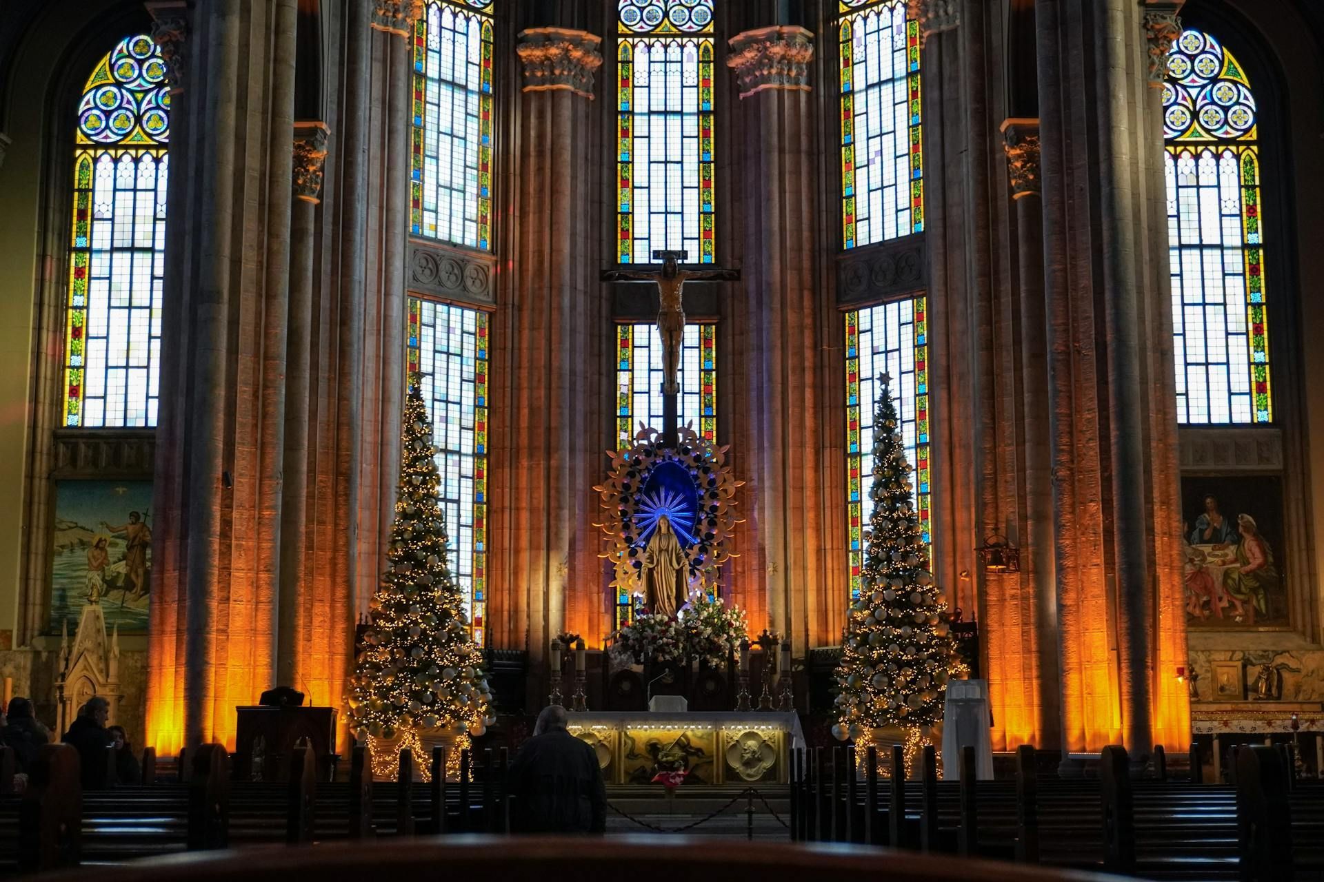 Inside a cathedral, two lit Christmas trees flank a central altar with a golden statue and a crucifix below stained glass.