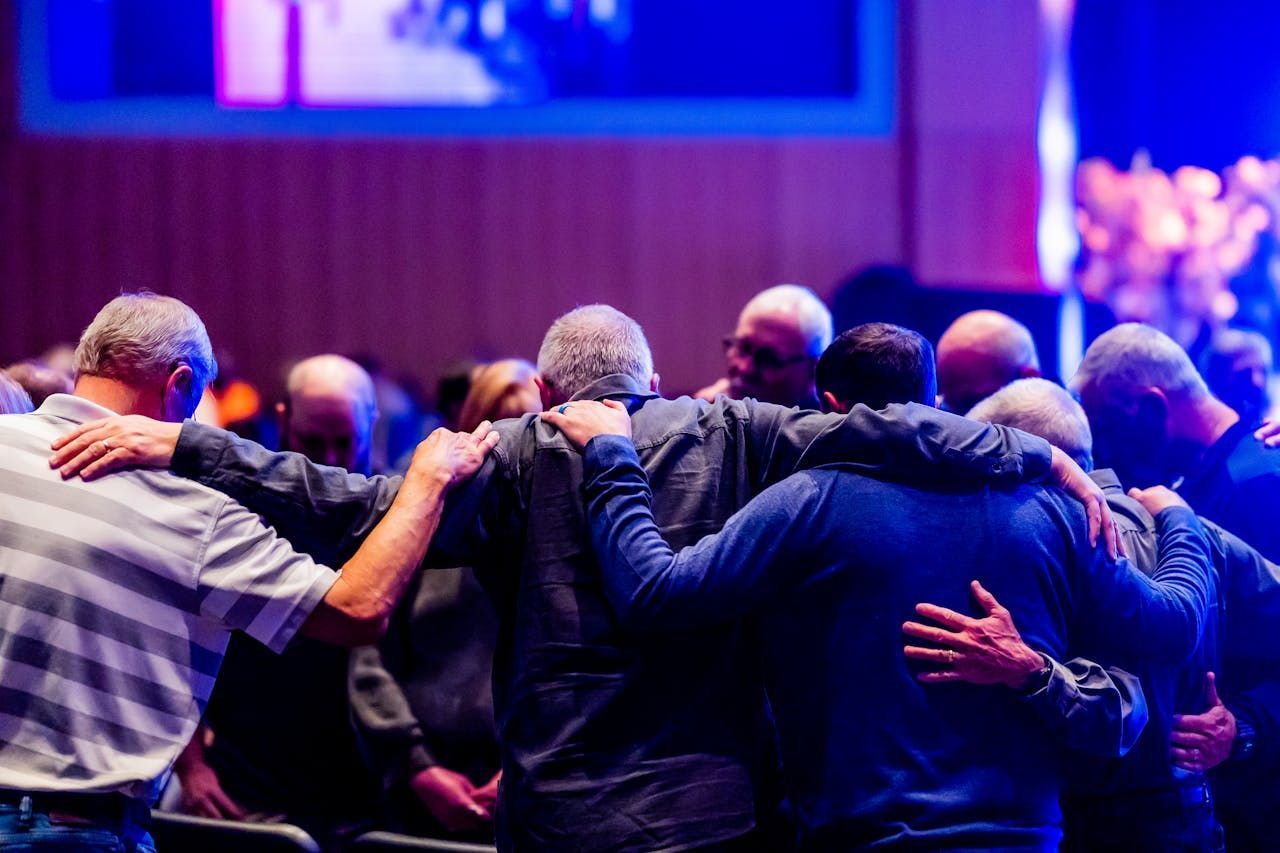 Men in a group hug, heads bowed, backs to camera. Lit stage in background, purple and blue hues.