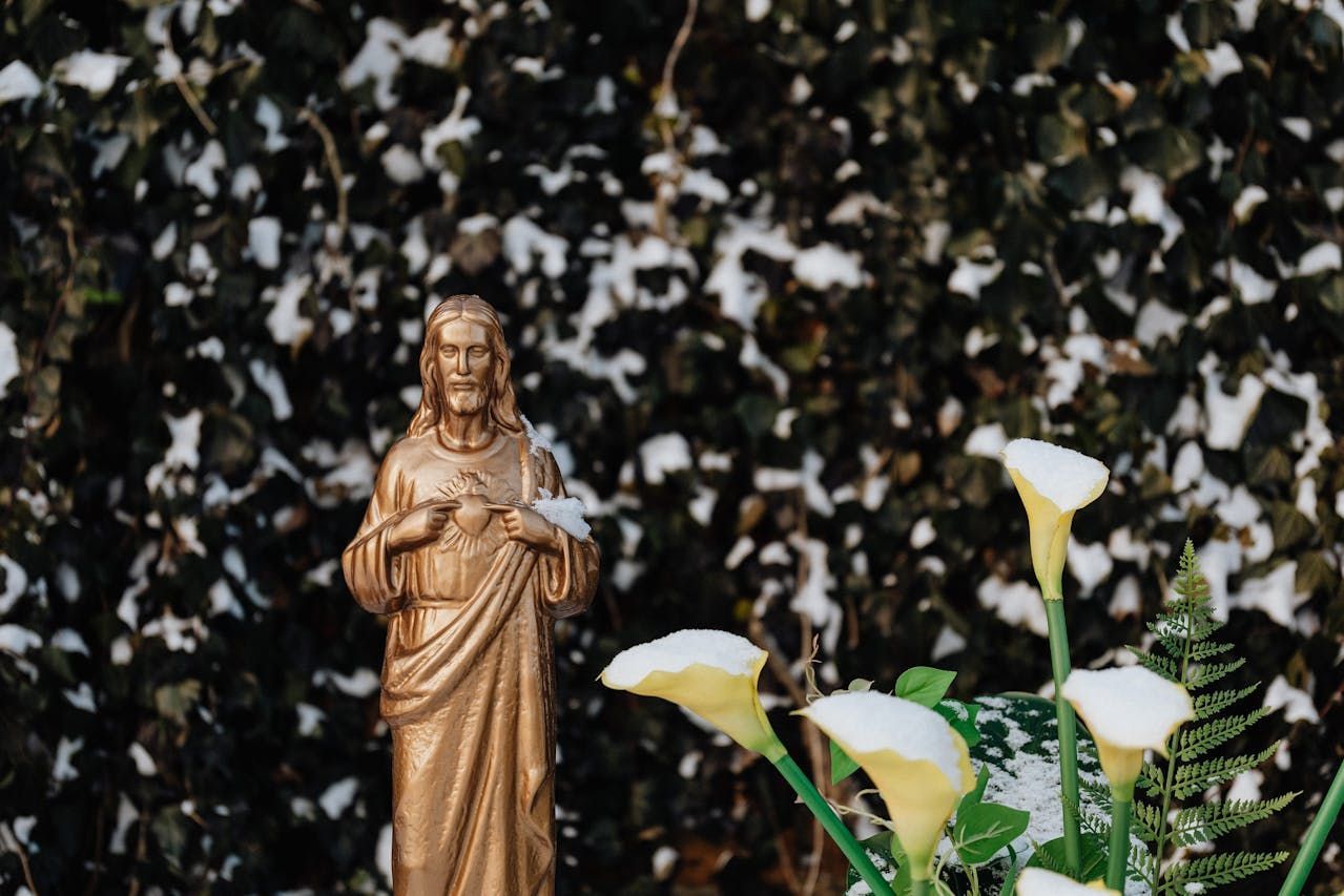 Golden statue of Jesus stands in snow-covered garden with calla lilies and leafy backdrop.