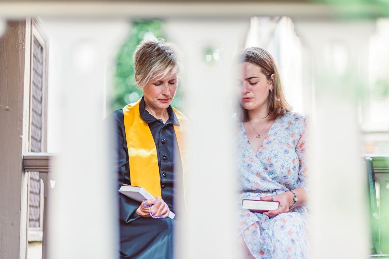 Woman with a yellow sash and woman in a floral dress sit on a porch, looking down at books.