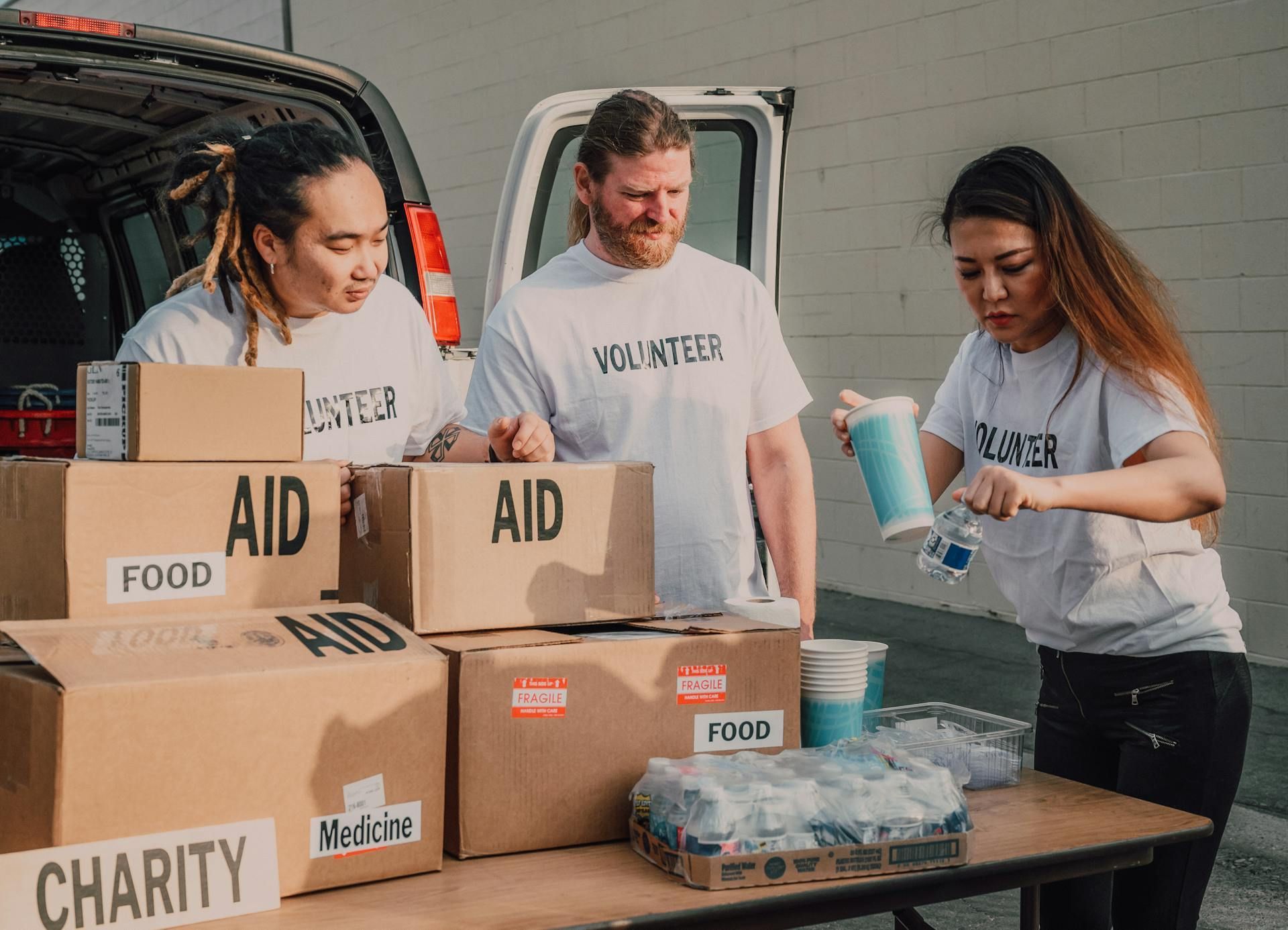 Three volunteers in matching white shirts work outside a van, organizing boxes labeled