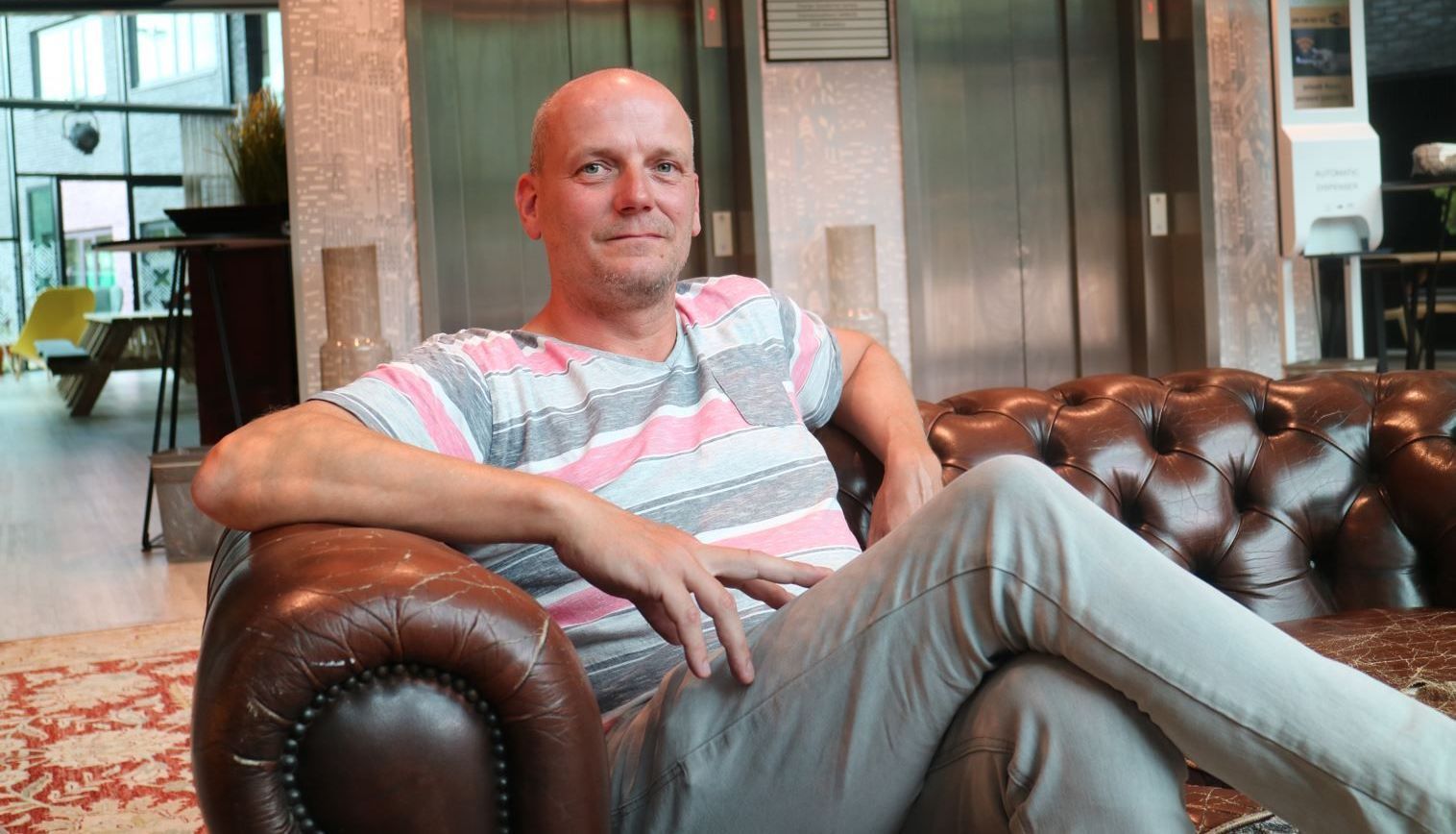 Een persoon ligt achterover op een bruine leren bank in een lichte lounge, gekleed in een gestreept shirt en een lichte broek.