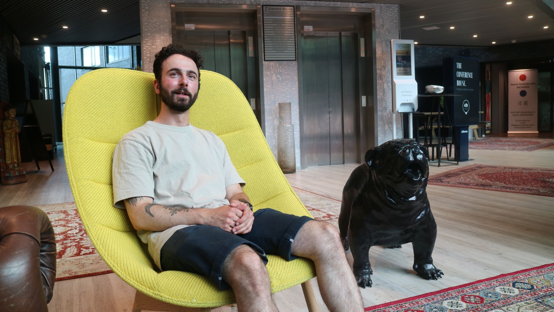 Een man zit op een gele stoel naast een zwarte hond in de lobby van een hotel.
