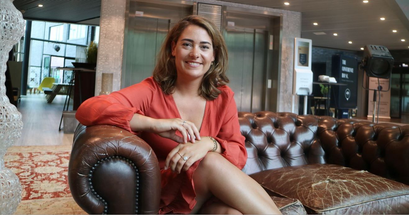 Vrouw in een oranje top zit op een bruine leren bank in een moderne lobby en lacht naar de camera.