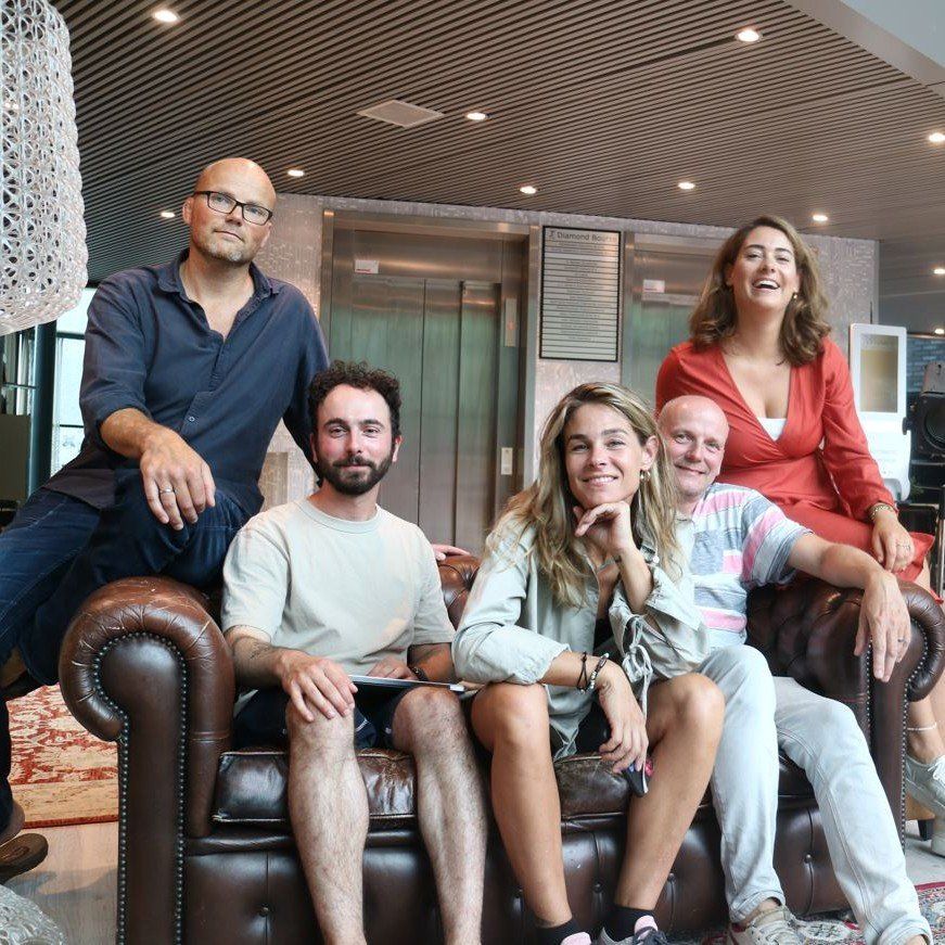 Vijf mensen zitten op een bruine leren bank in een moderne kantoorlounge en lachen naar de camera.