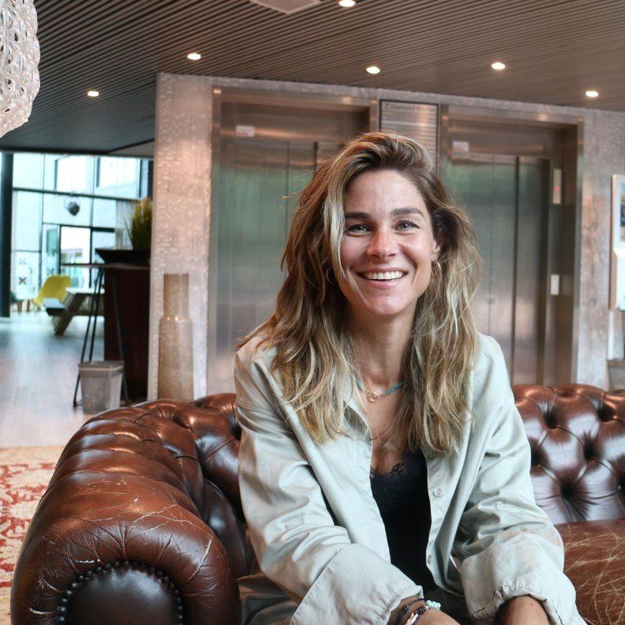 Een glimlachende vrouw zit op een bruine leren bank in een moderne lobby met liften op de achtergrond.