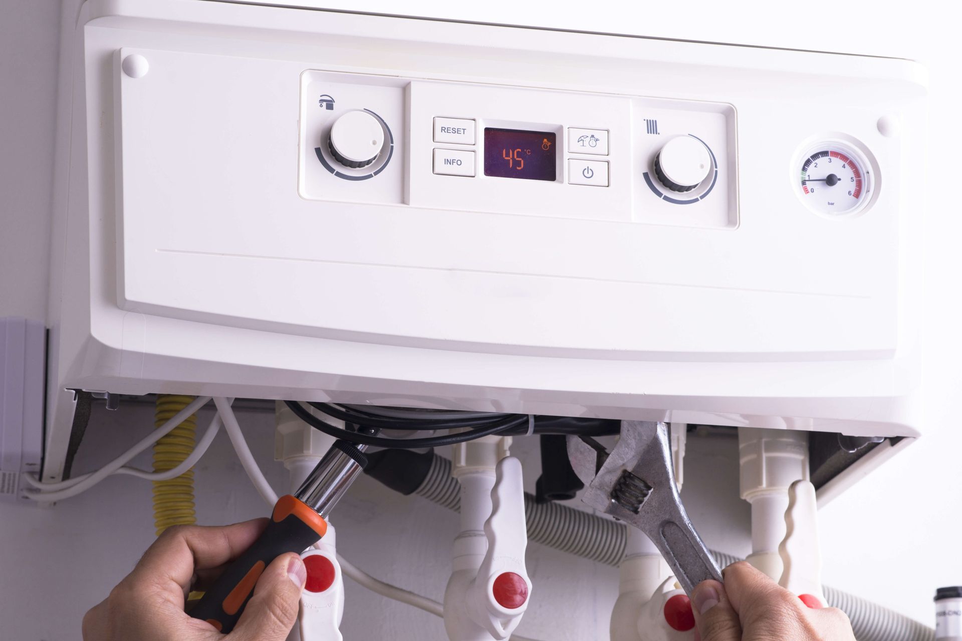 Person tightening pipes under a white boiler control panel with a screwdriver