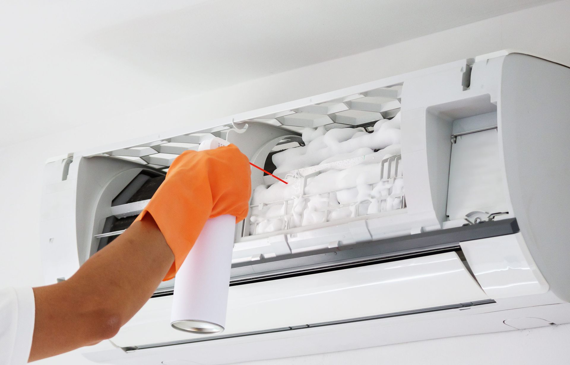 Person in orange gloves cleaning the inside of a wall-mounted air conditioner with a spray can.