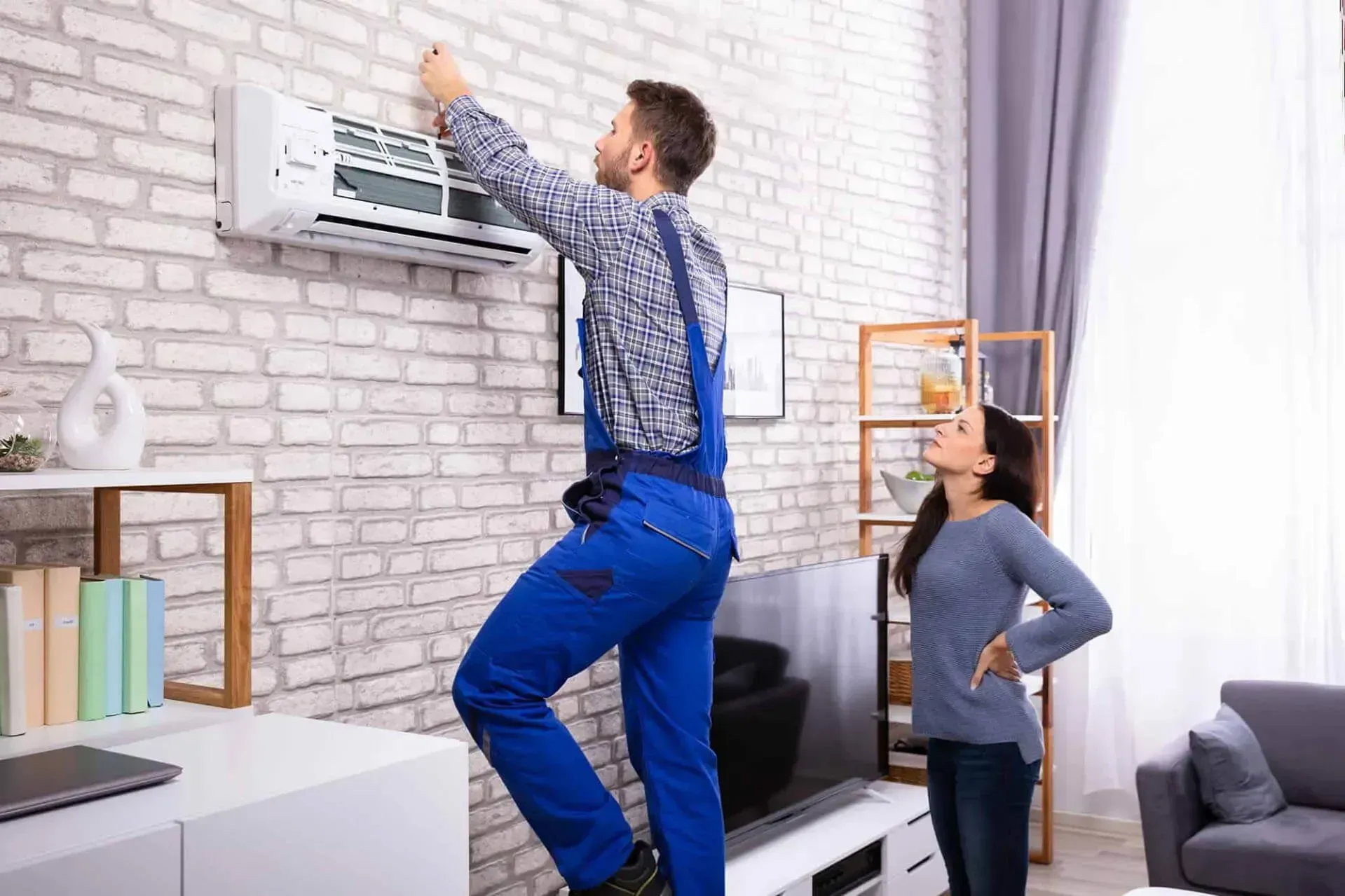 Two people adjusting a wall-mounted air conditioner in a bright living room