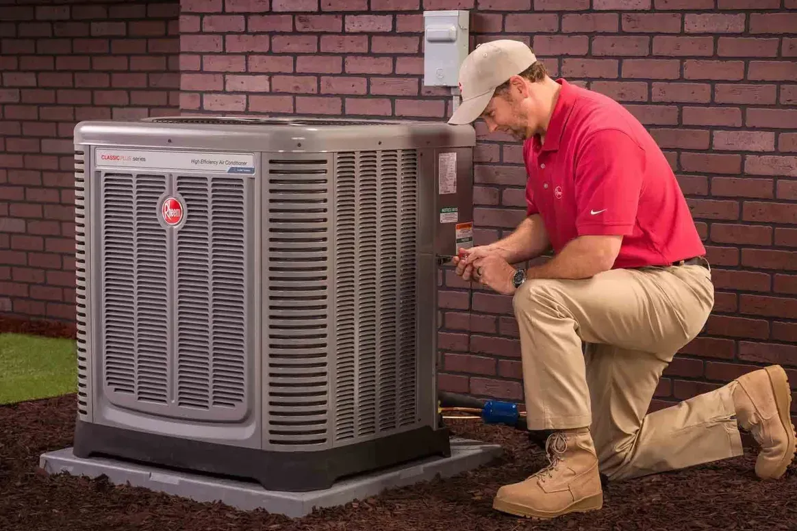 Technician in red shirt kneels beside outdoor HVAC unit, inspecting it against a brick wall.