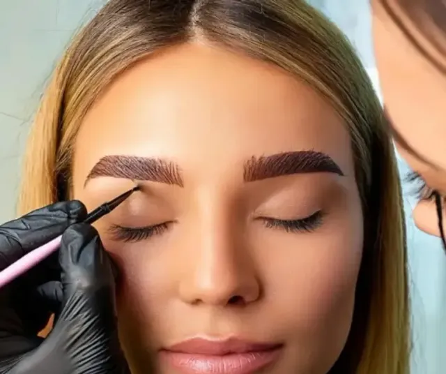 Gloved hands shaping a woman’s eyebrow with a brow pencil, eyes closed