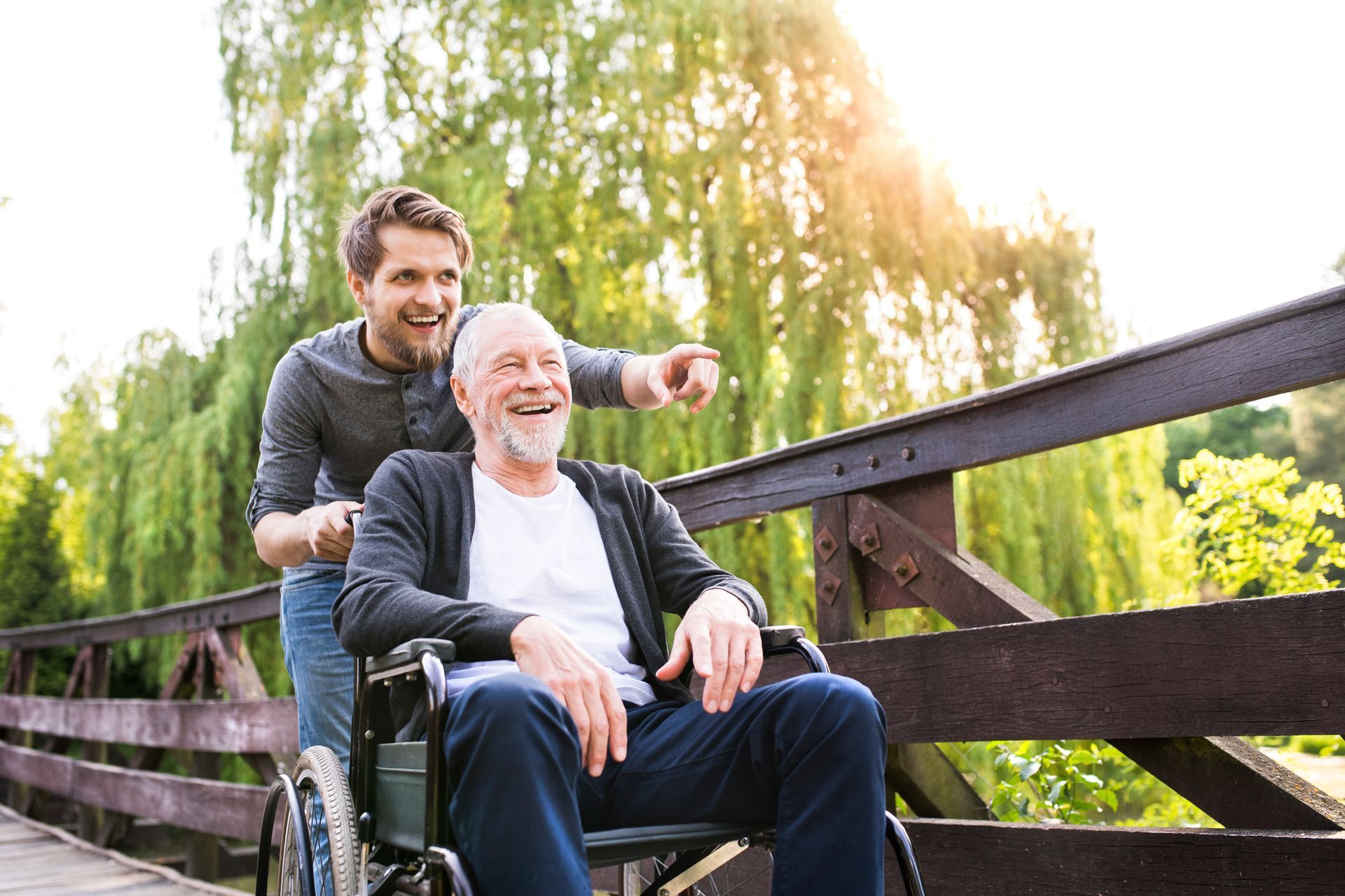 Man pushing another man in wheelchair across a wooden bridge outdoors, both smiling.