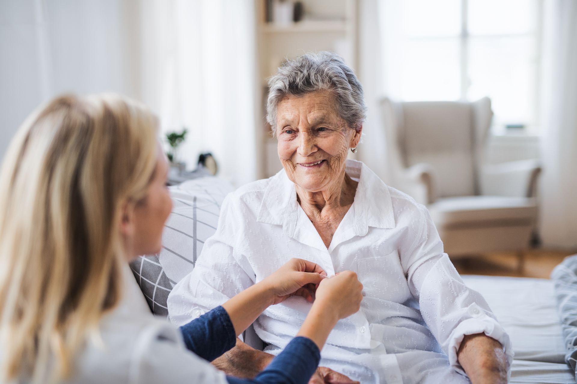 Blonde caregiver buttons a white shirt for an older woman seated indoors, smiling.
