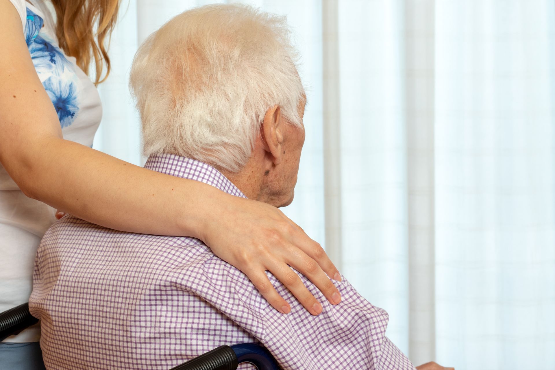 Person with white hair in a wheelchair looks out the window; a person's hand is on their shoulder.