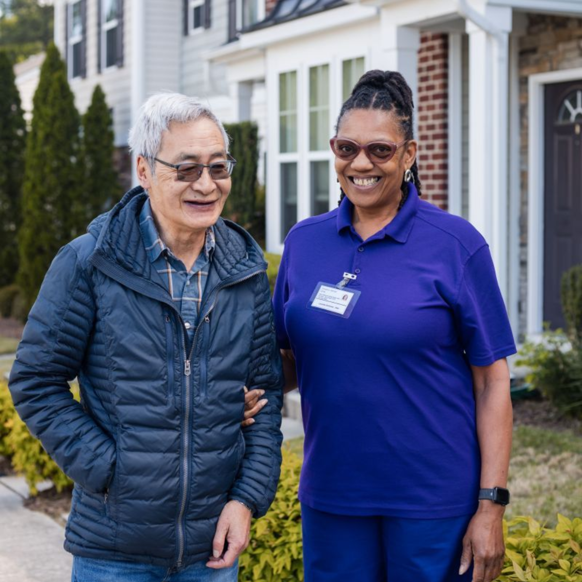Man with glasses and woman in purple shirt walking outside a house; woman is helping the man walk.
