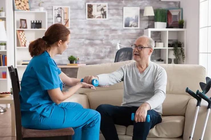 Healthcare worker assists a man with arm exercises using weights; seated on a sofa.